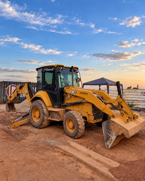 Retroescavadeira CAT 416F2 com a marca CARMAC na cabine, em canteiro de obra ao entardecer
